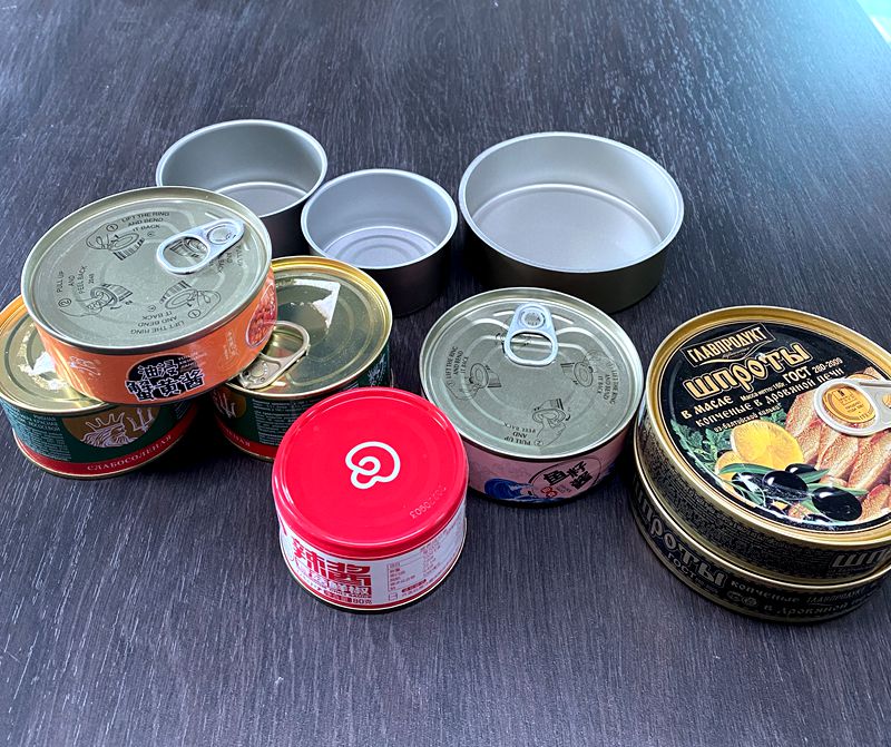Assortment of metal canned foods on a dark table; several lids removed and pull-tabs visible.